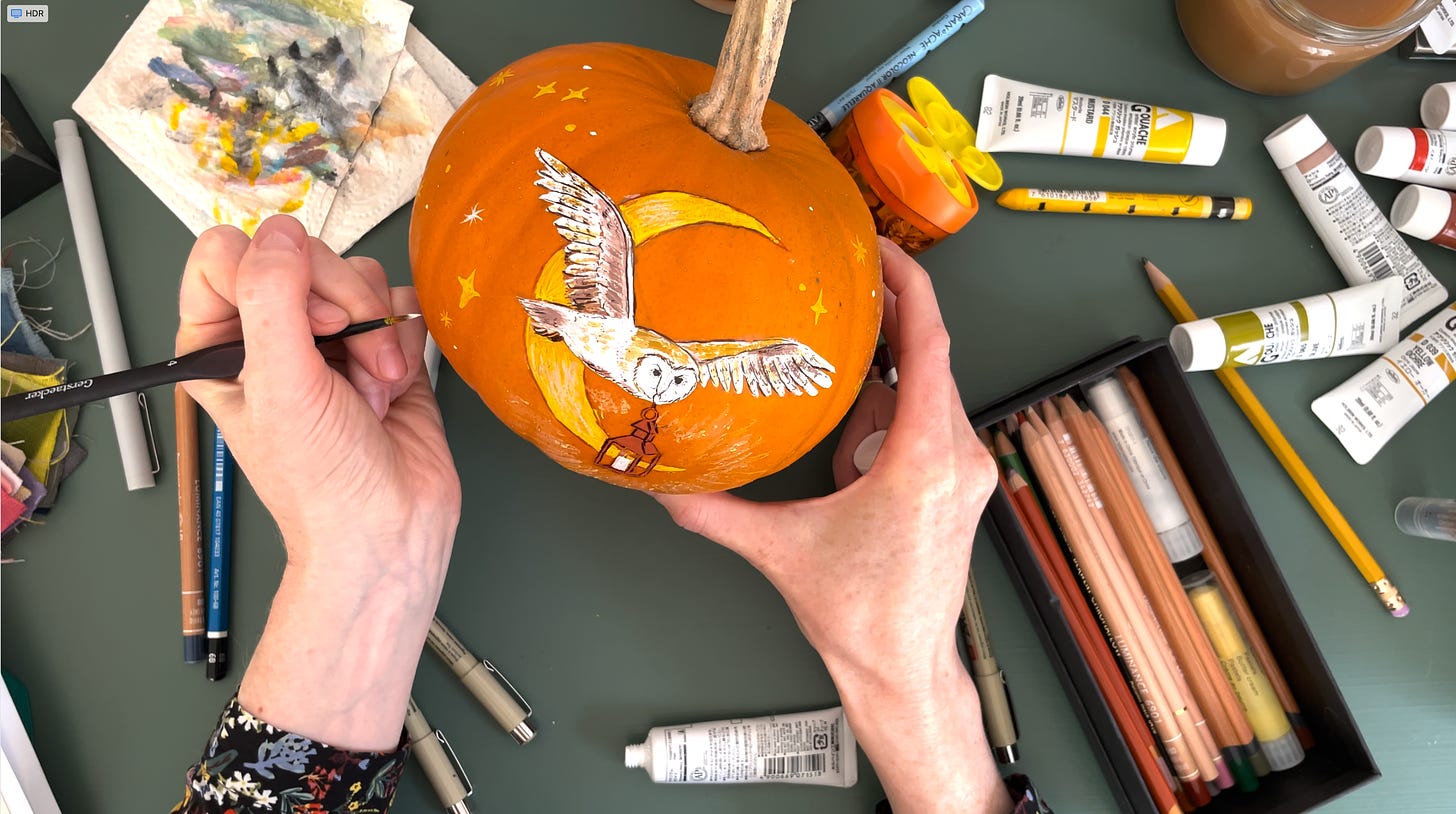 overview of a desk, 2 hands painting an owl and a moon on an orange pumpkin with art materials around it