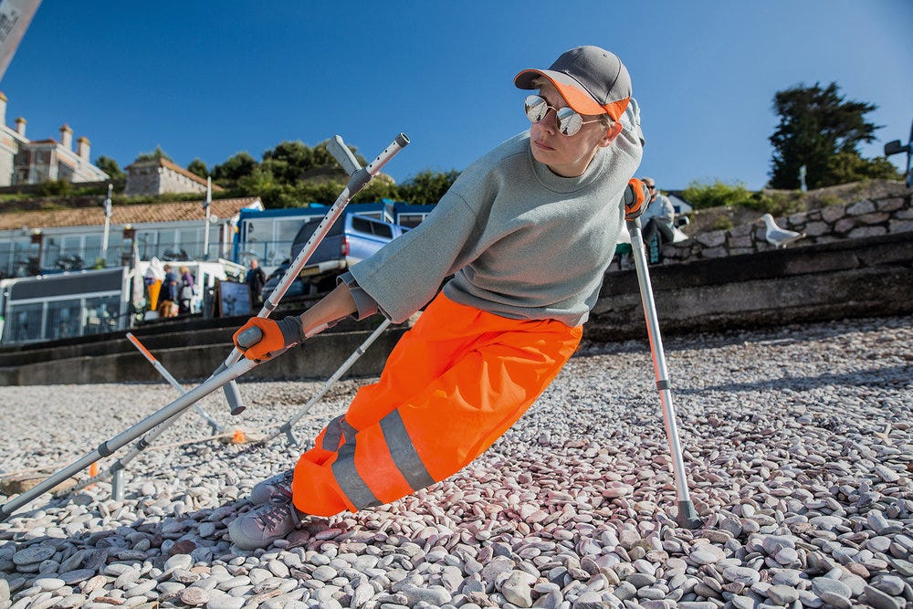 A person is on a stony beach on a cloudless day, leaning deeply to one side with metal crutches. They are wearing bright neon orange pants and grey clothing with pops of the same orange.