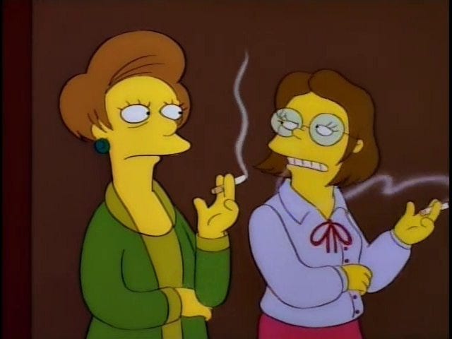 Screencap from The Simpsons. Edna Krabappel and Mrs. Hoover smoke off to the side of a presentation and discuss the word Embiggens.