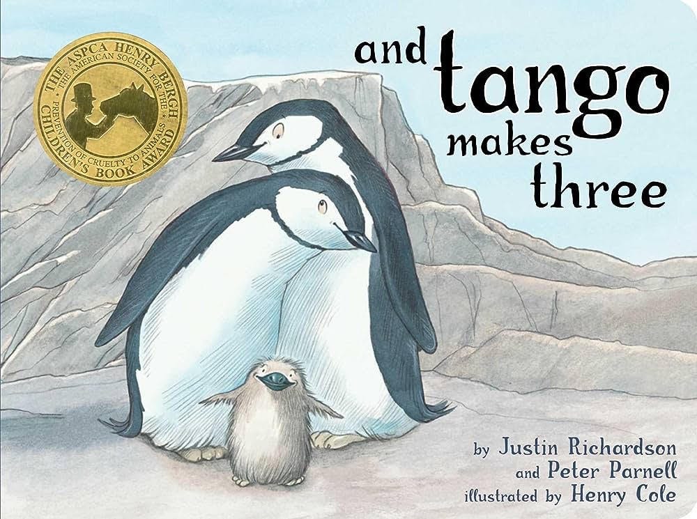 The photo is a cover photo of a children's picture book. The book cover includes an illustration of two adult penguins sheltering a baby penguin as they all sit upon boulders. The text reads, "And Tango Makes Three by Justin Richardson and Peter Parnell, illustrated by Henry Cole". 
