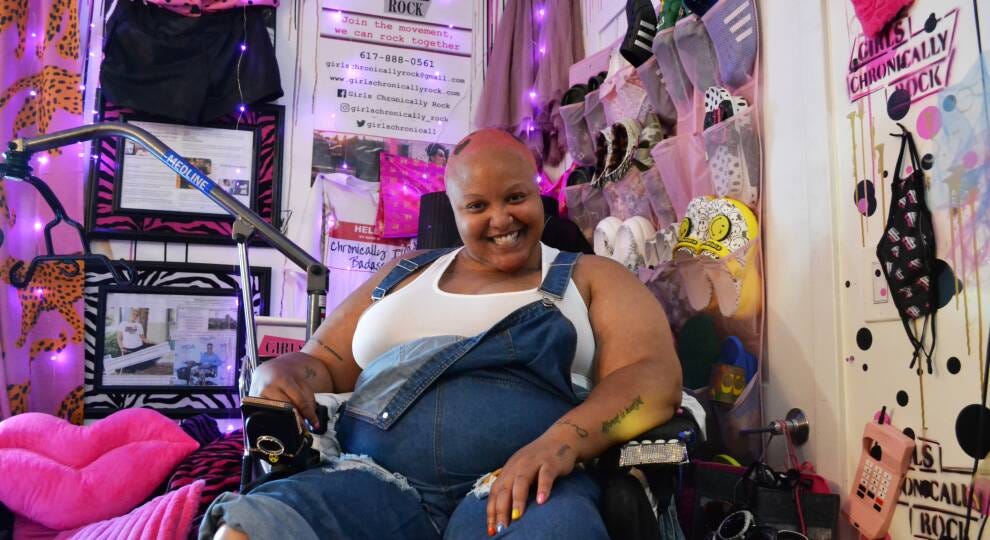 A woman in a wheelchair wears jean overalls with short pink cropped hair. She smiles at the camera. She is surrounded by vibrant clothing and decor, mostly pink, black and animal print. 