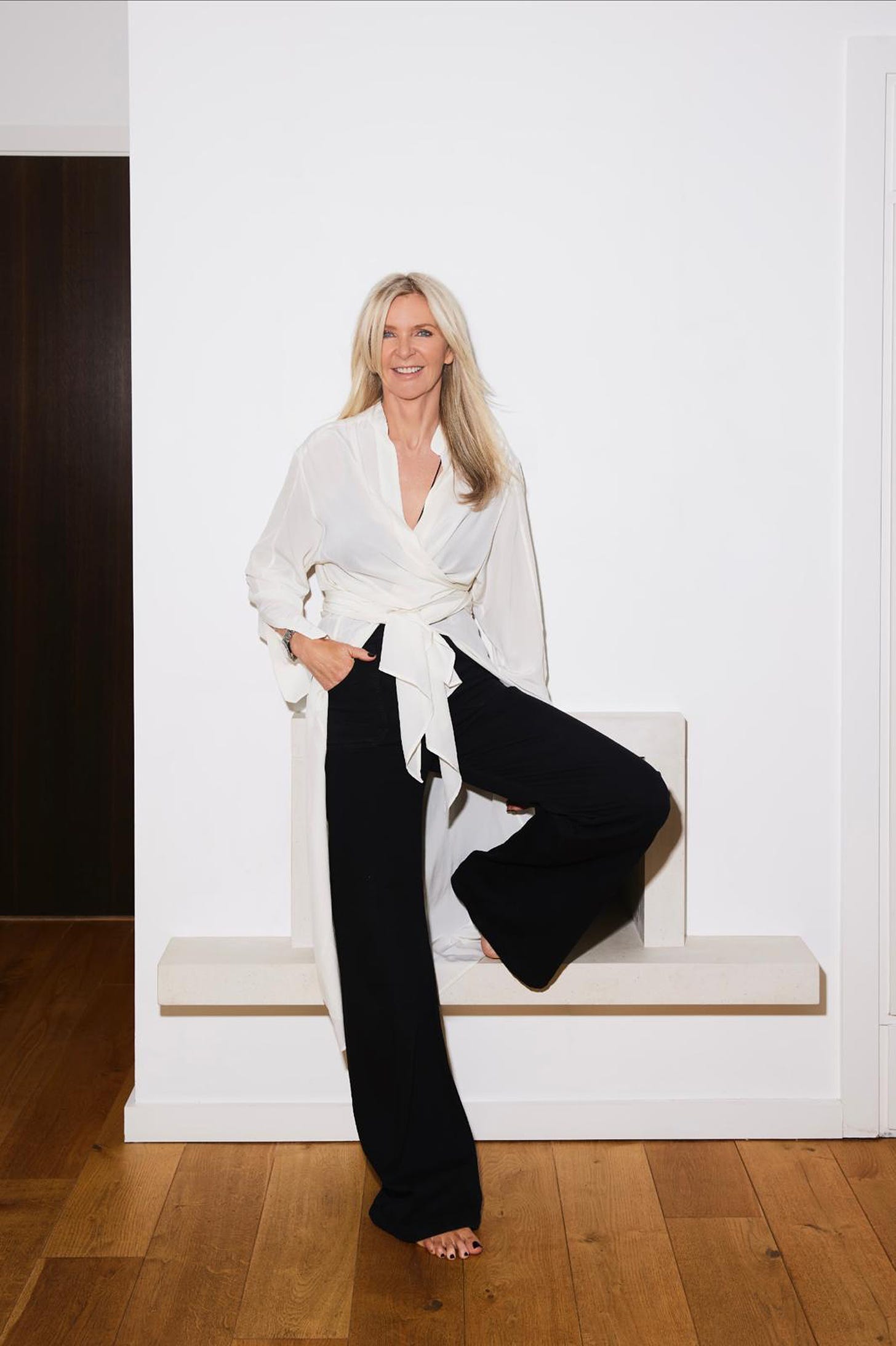 Amanda Wakeley standing against a white wall, wearing black wide legged trousers and a white shirt, tied at the waistline