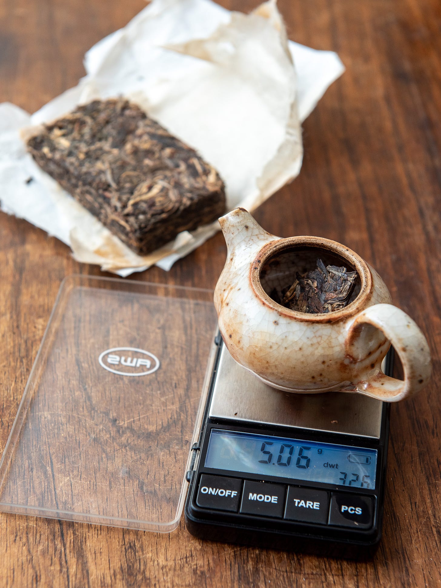 ID: Teapot on gram scale showing 5 grams of tea, with brick in background
