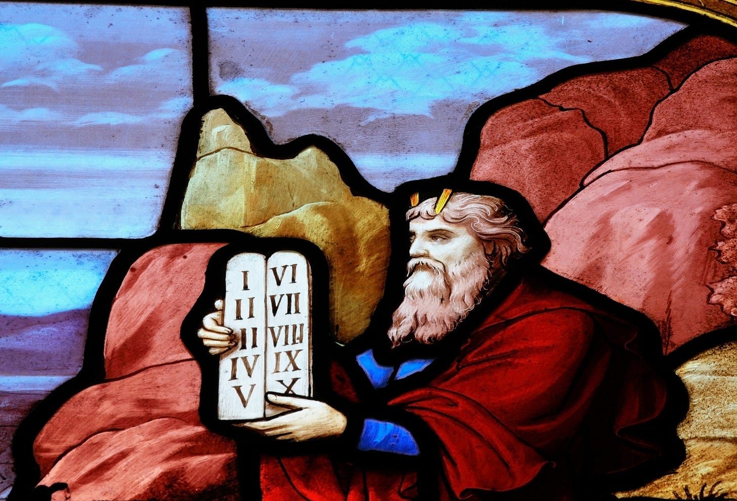 Image depicts Moses holding tablets with ten Roman numberals. Rocks and sky in background.