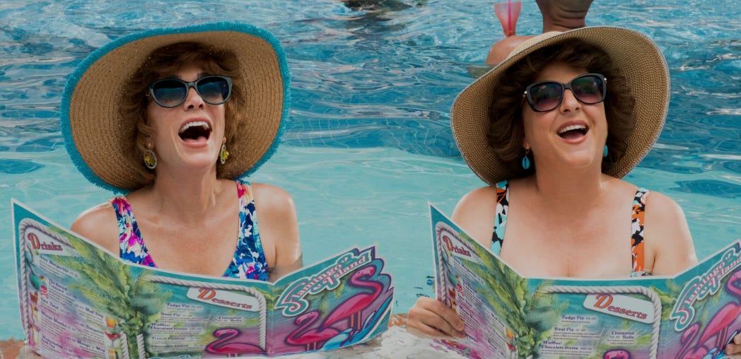 Barb and Star smiling up from their place in the pool, both holding huge resort menus and wearing floppy hats and sunglasses