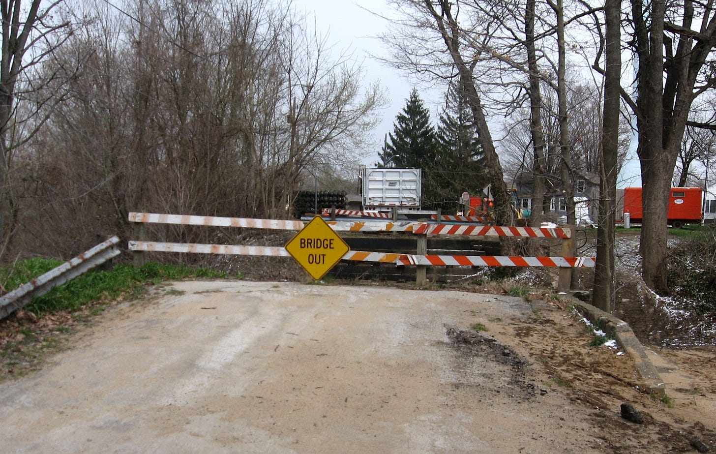 A dirt road leads up to an orange-and-white road block with a yellow sign on it reading "BRIDGE OUT." There are trees without leaves in the background and the sky is gray.