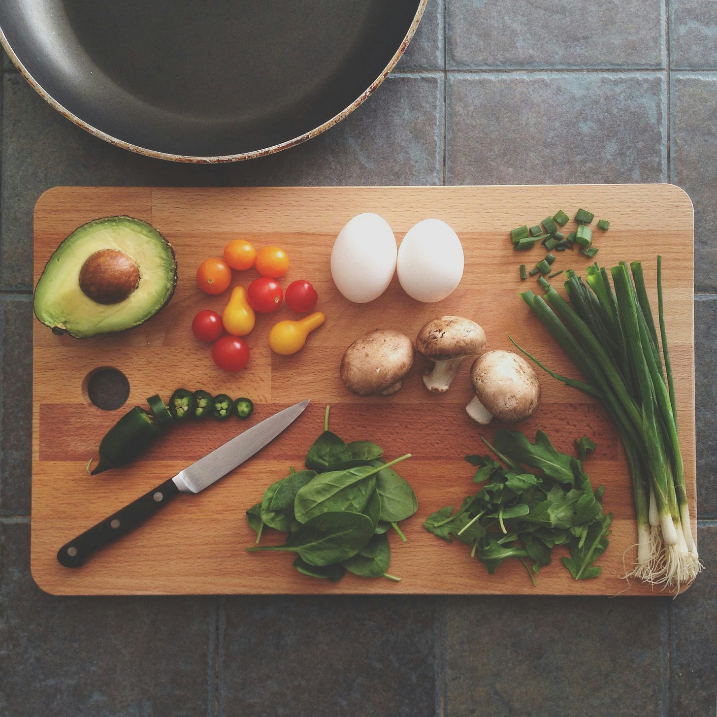 top view of a wooden cutting board on a stone tiled surface, with a frying pan partially visible above it. a knife with a black handle and many different sliced foods sit on the cutting board - avocado, tomatoes, mushrooms, spinnach, green onion