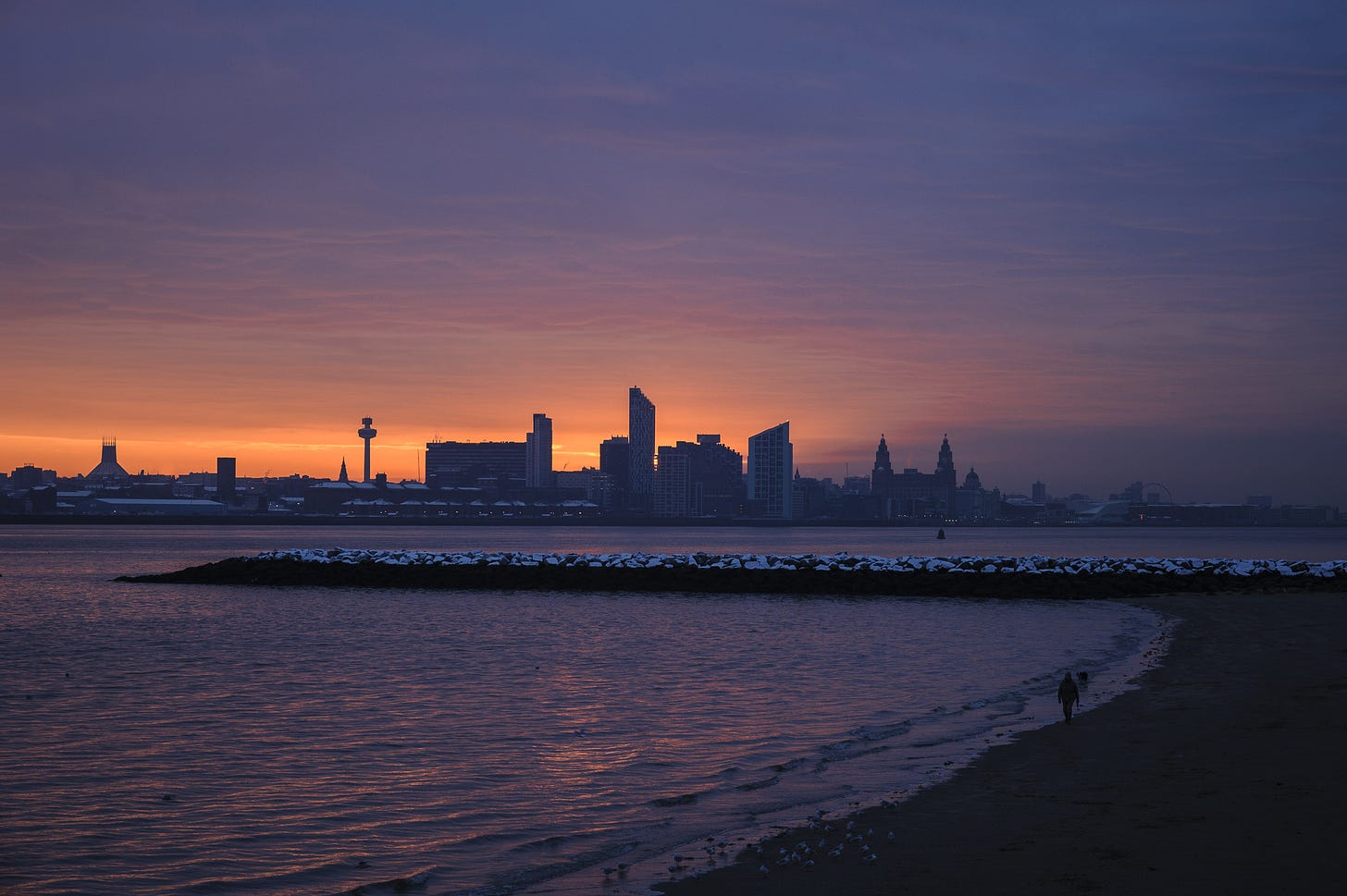As the sky turns purple a person walks along the river bed of the Mersey while the tide is out. It’s a cold morning and there is a dusting of snow on the rocks.