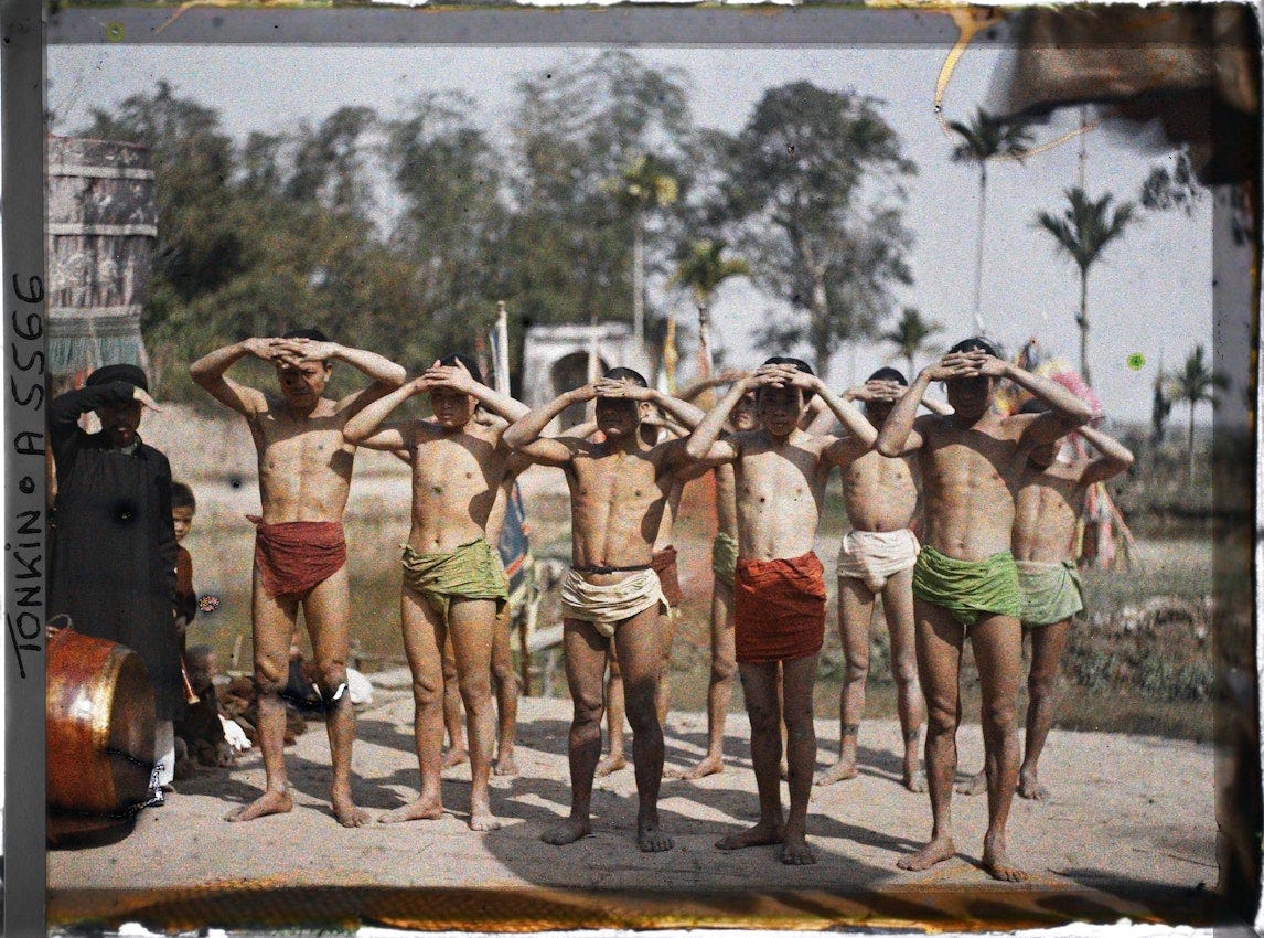 Group of young boys wearing colored loincloths standing in formation with hands raised to shade their eyes from sun, supervised by figure in black clothing, with tropical landscape and palm trees behind them in outdoor setting.