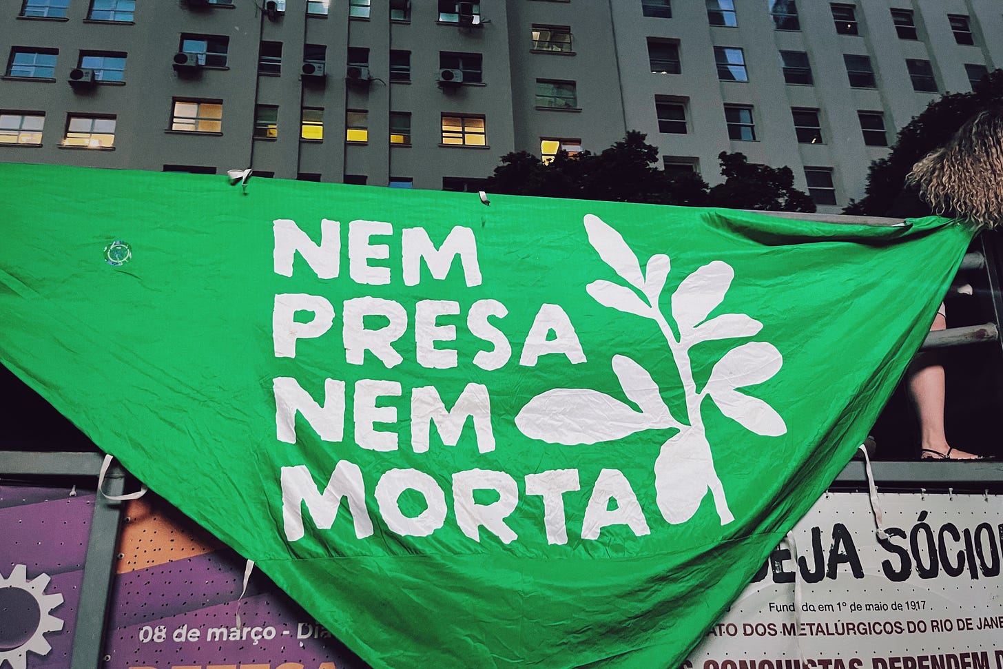 Photograph of a large green banner that says NEM PRESA NEM MORTA, hanging from the side of a bus during the Women's Day march in Rio de Janeiro.