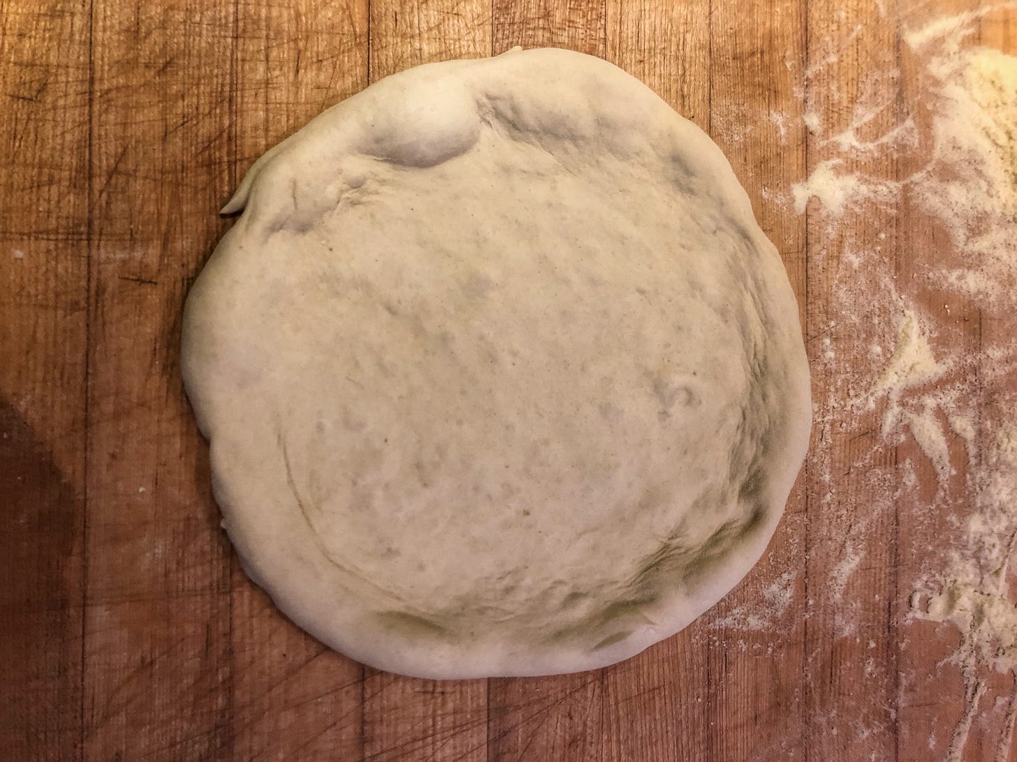 A disk of partially-stretched pizza dough on a wooden counter that is dusted with semolina. 