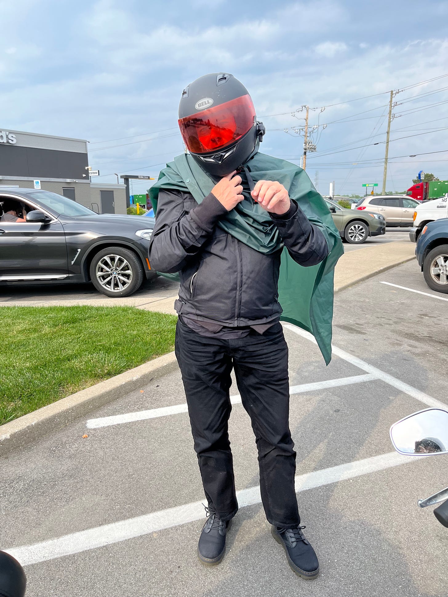 Derrick stands in a parking lot with his helmet still on, putting a rain cover/coat over himself.