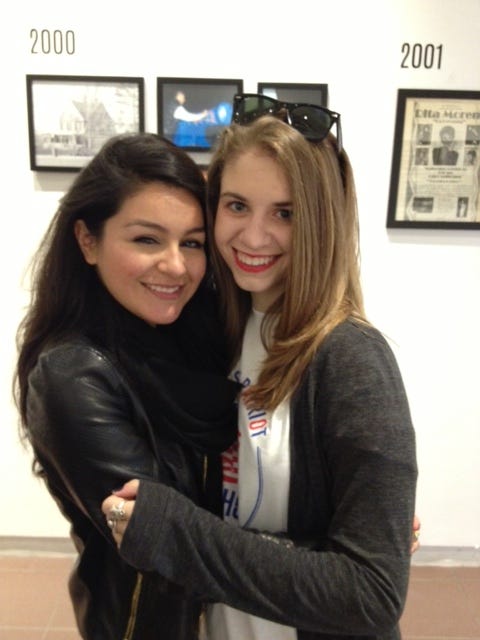 Two woman with long hair standing next to each other with their arms around each other. They are in front of a white wall with pictures on it. 