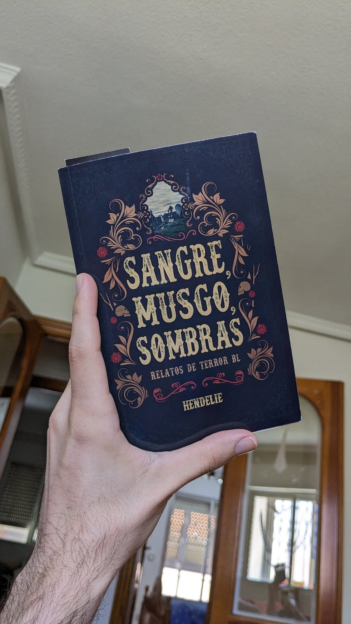 Una foto del libro de Sangre, Musgo, Sombras, sujetada por una mano izquierda, detrás se ve el techo blanco y una puerta con cristalera abierta. La portada del libro tiene el título en el centro, el subtítulo "relatos de terror BL" justo debajo en pequeño, todo está envuelto en varias cenefas de colores cálidos con motivos vegetales y calaveras. En la parte superior, hay una pequeña imagen de un cementerio. 