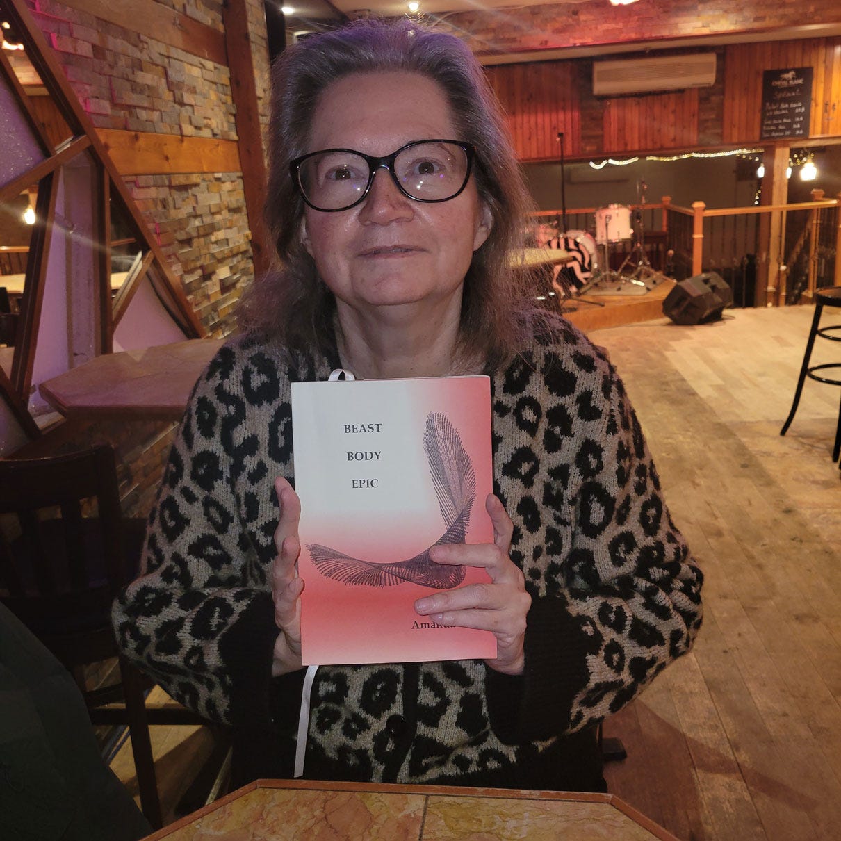 Amanda wears a leopard print sweater and holds a copy of Beast Body Epic at a pub in Montreal