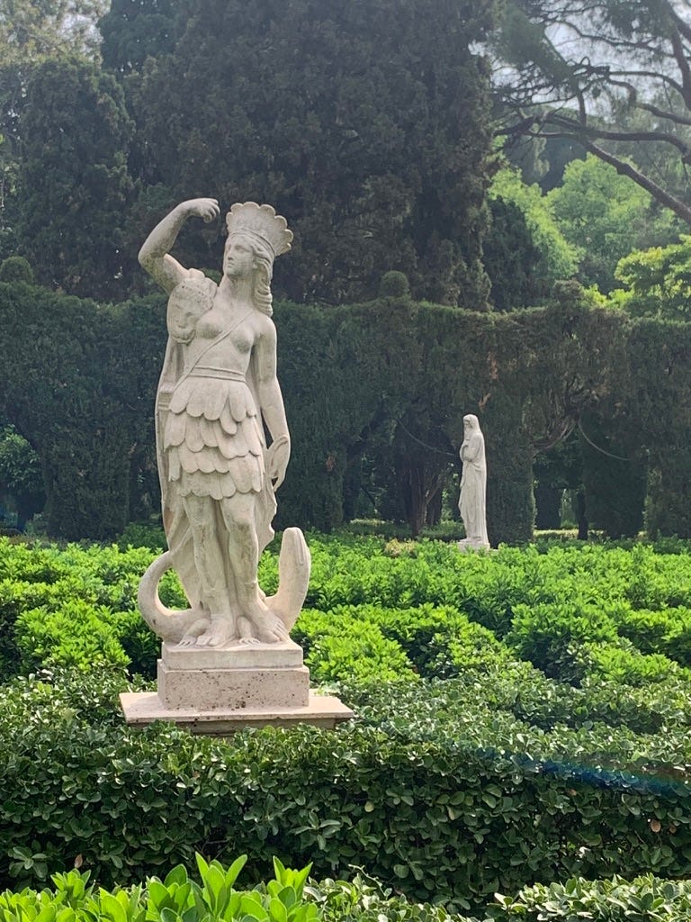 hedges and trees, with a white stone statue of a woman in a feather skirt and headdress, with an 'alligator' at her feet