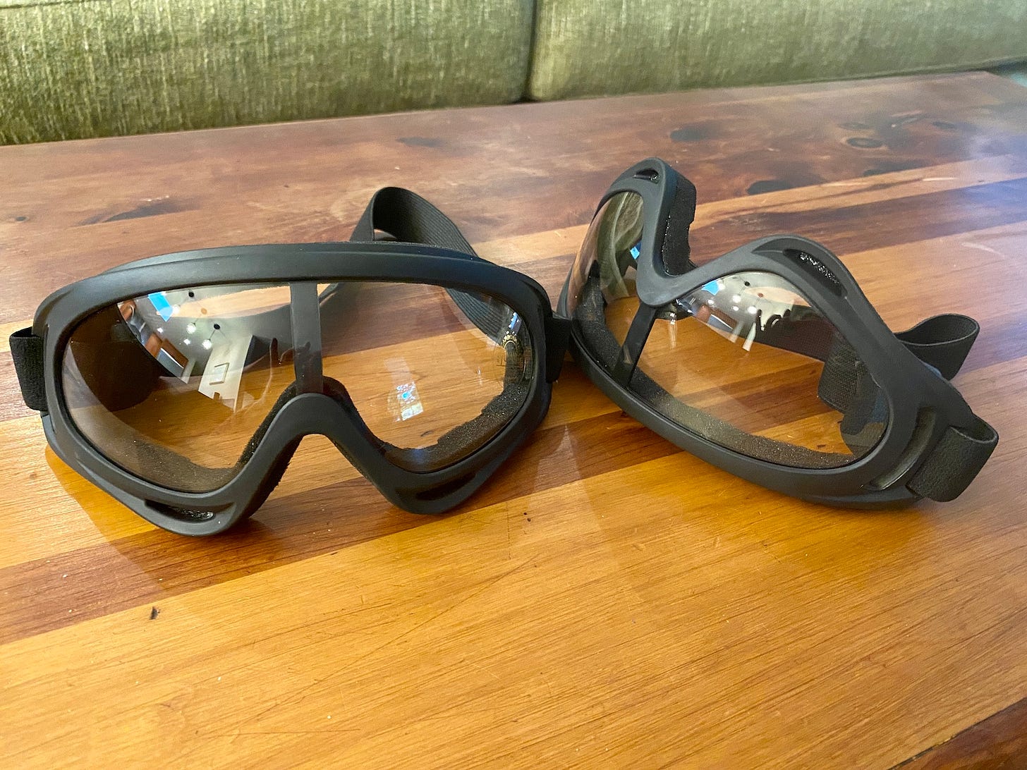 A pair of ski goggles with black frames and clear lenses sits on a wooden tabletop.