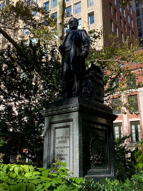 A dashing statue of Chester A Arthur in Madison Square. He stands in front of an ornate chair, his right hand clenched in front of him.