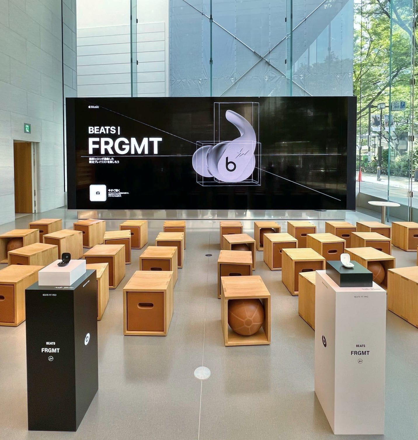 Fragment Design Special Edition Beats Fit Pro displayed at Apple Omotesando. The earbuds are placed on two black and white pedestals in the Forum. A matching graphic is displayed on the video wall.