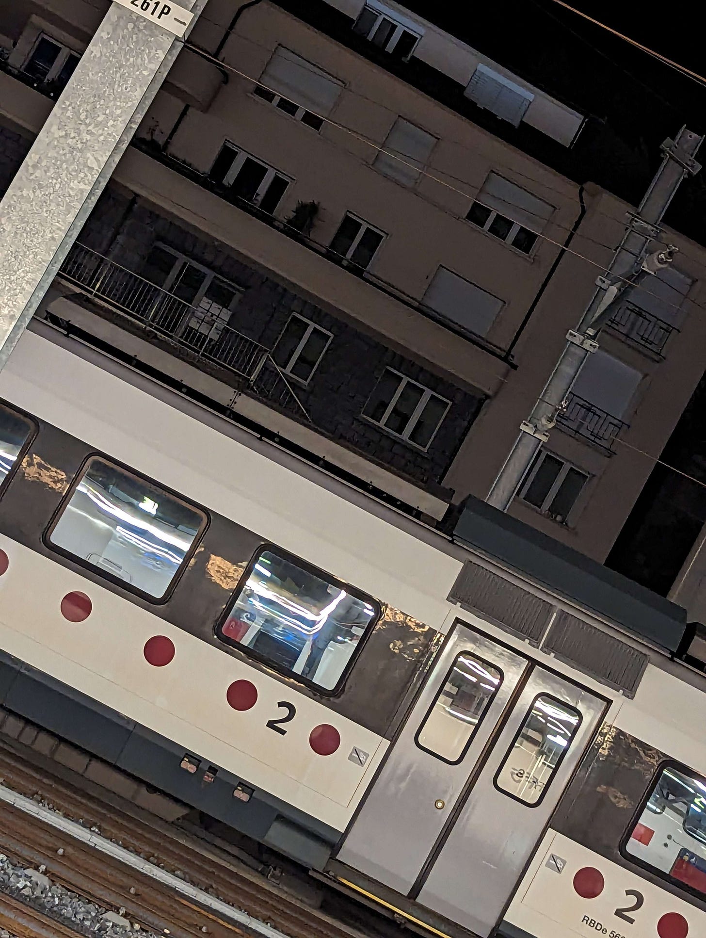 A white train with red dots lining its side sits on train tracks in front of a beige residential building.