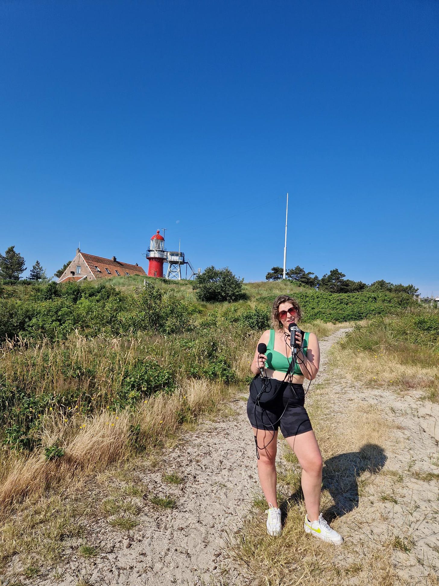 oto van Marie-Lotte, een vrouw van eind 30 met bruin haar met blonde highlights en een zonnebril op. Ze heeft een groen topje aan en een zwarte korte broek. Ze staat op een duin, in de achtergrond zie je de rode vuurtoren van Vlieland en de lucht is strakblauw. In haar hand heeft ze een recorder met daaraan twee microfoons. Ze houdt een van de microfoons voor haar mond en praat er in.