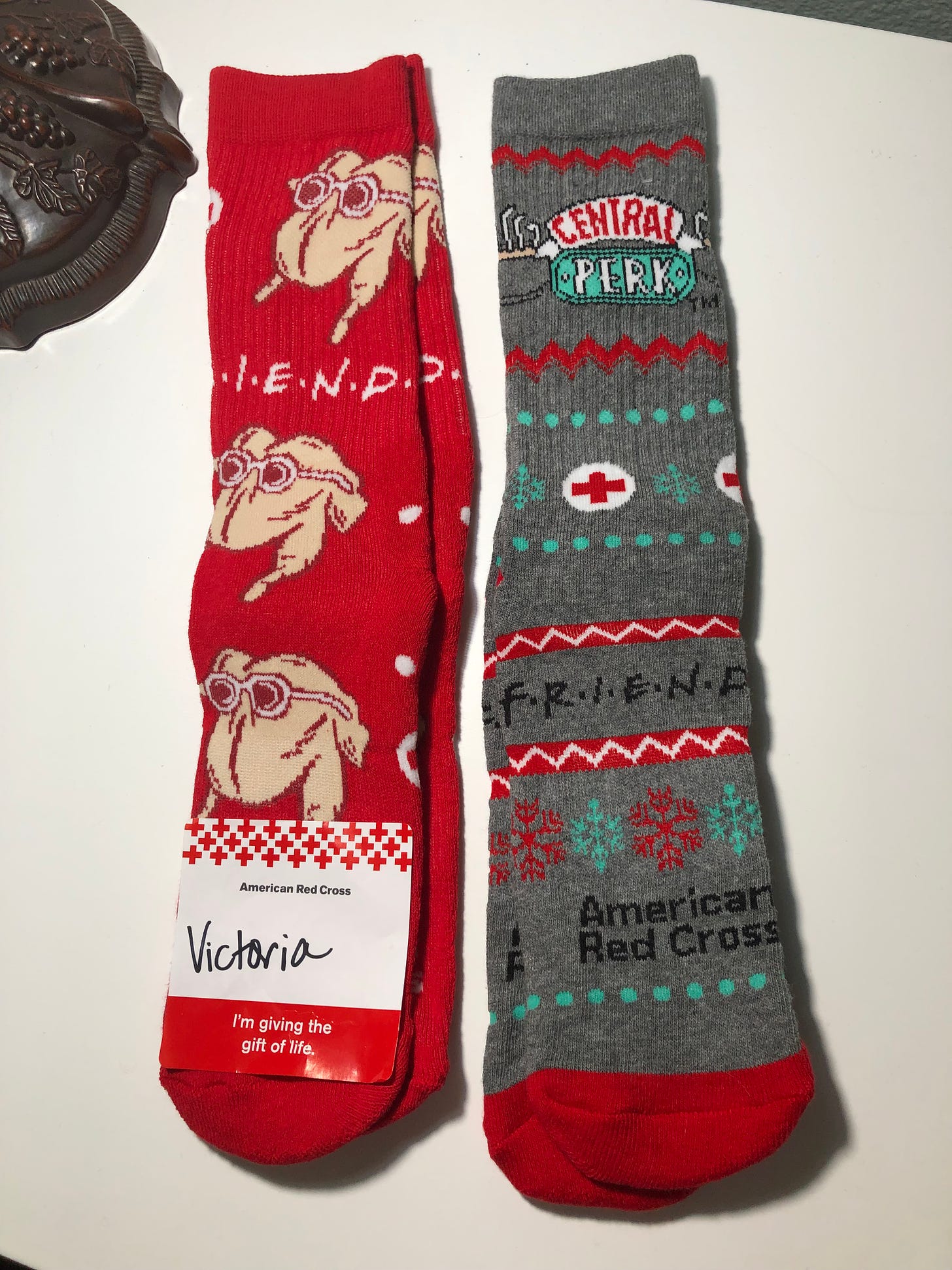 Two pair of socks. One is red and has a turkey prepared for cooking, wearing sunglasses and the word “Friends.” The other is gray and has snowflakes, the logo of teh American Red Cross, and the word “Friends.”