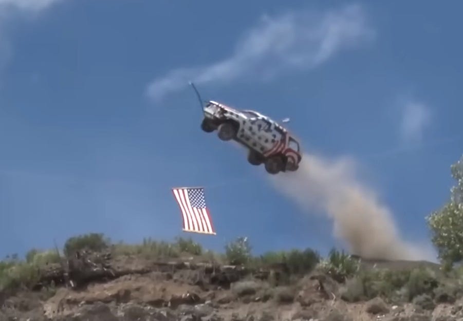 Video screenshot of a driverless, star-spangled SUV zooming off a cliff, trailing dust, with a US flag suspended by wires near the jump site. 1320video on YouTube