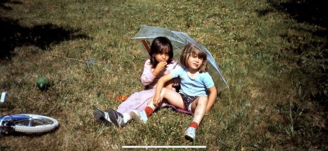 Two 5 year old kids sitting on the grass in nature on a sunny day under an umbrella. The kid to the left is white with semi long brown hair and is dressed in a pink sweat suit. She is looking shyly at the camera with her legs straight and next to each other and her right hand in her mouth. The kid to the right (me) is white with a light brown bob cut and is looking defiantly at the camera with her legs wide open. There is a blue and white bicycle wheel on the left side of the frame. 