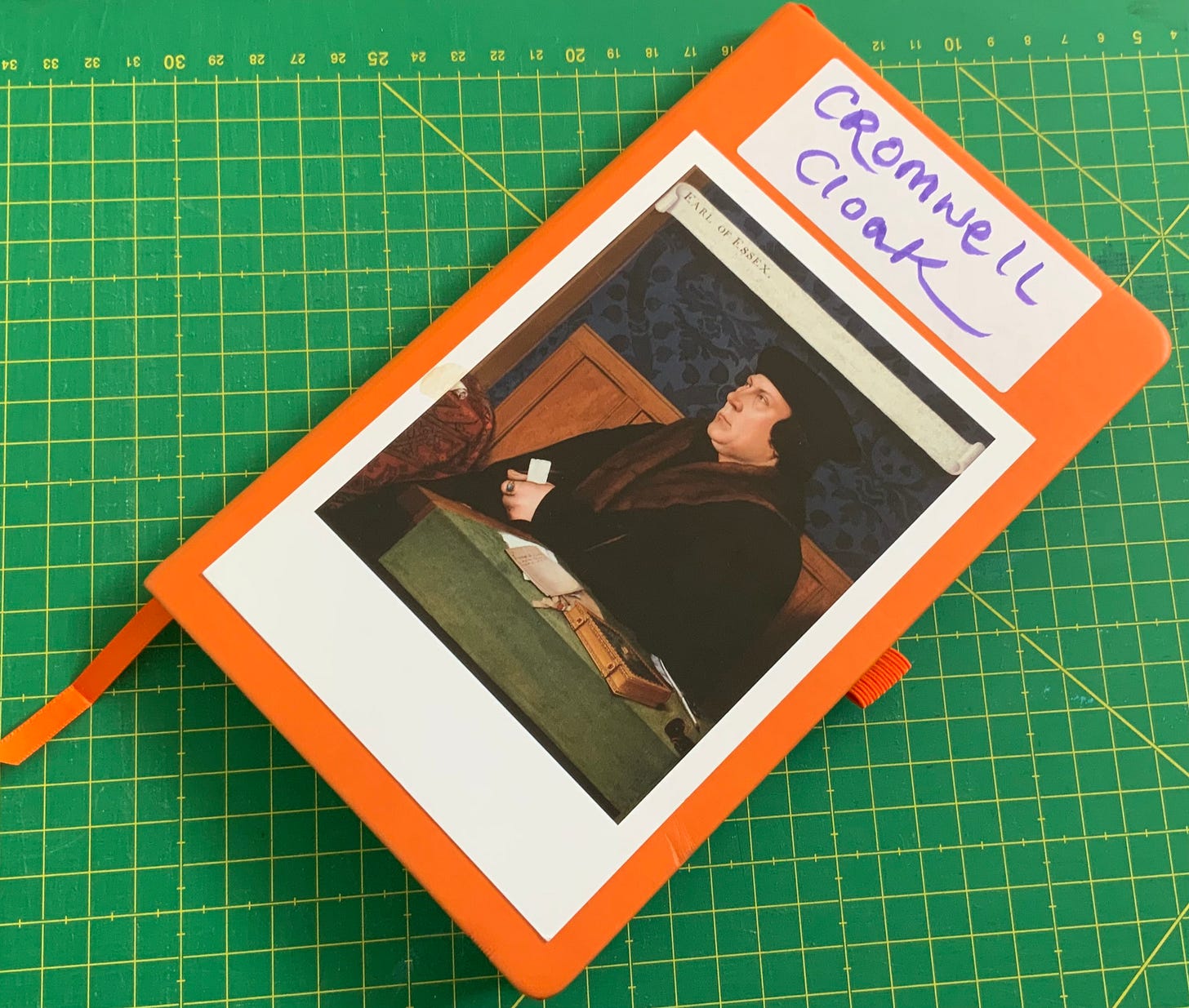 An orange notebook with a postcard of Thomas Cromwell stuck to the front, with a label “Cromwell Cloak”.