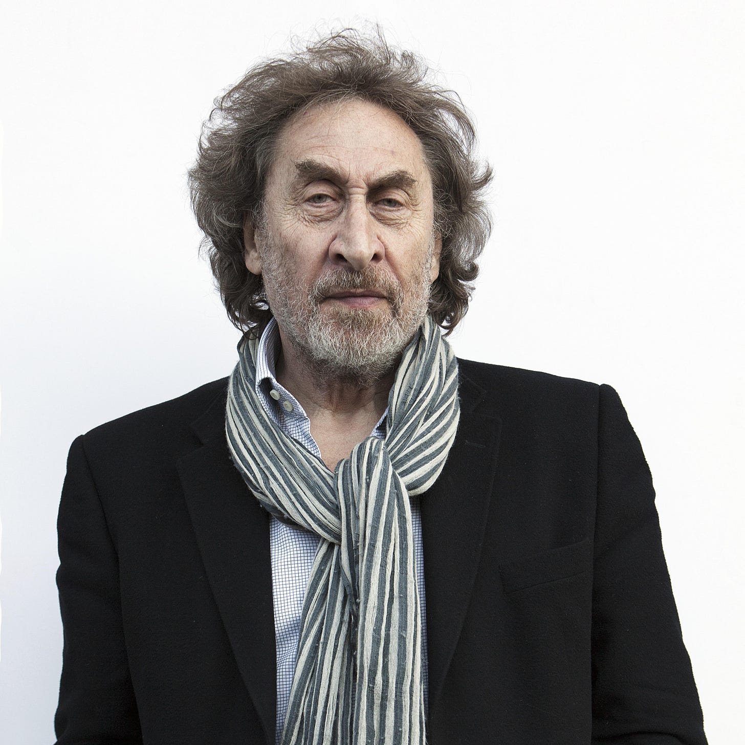 Older white man with short gray beard and wild brown hair wearing a blazer and scarf.