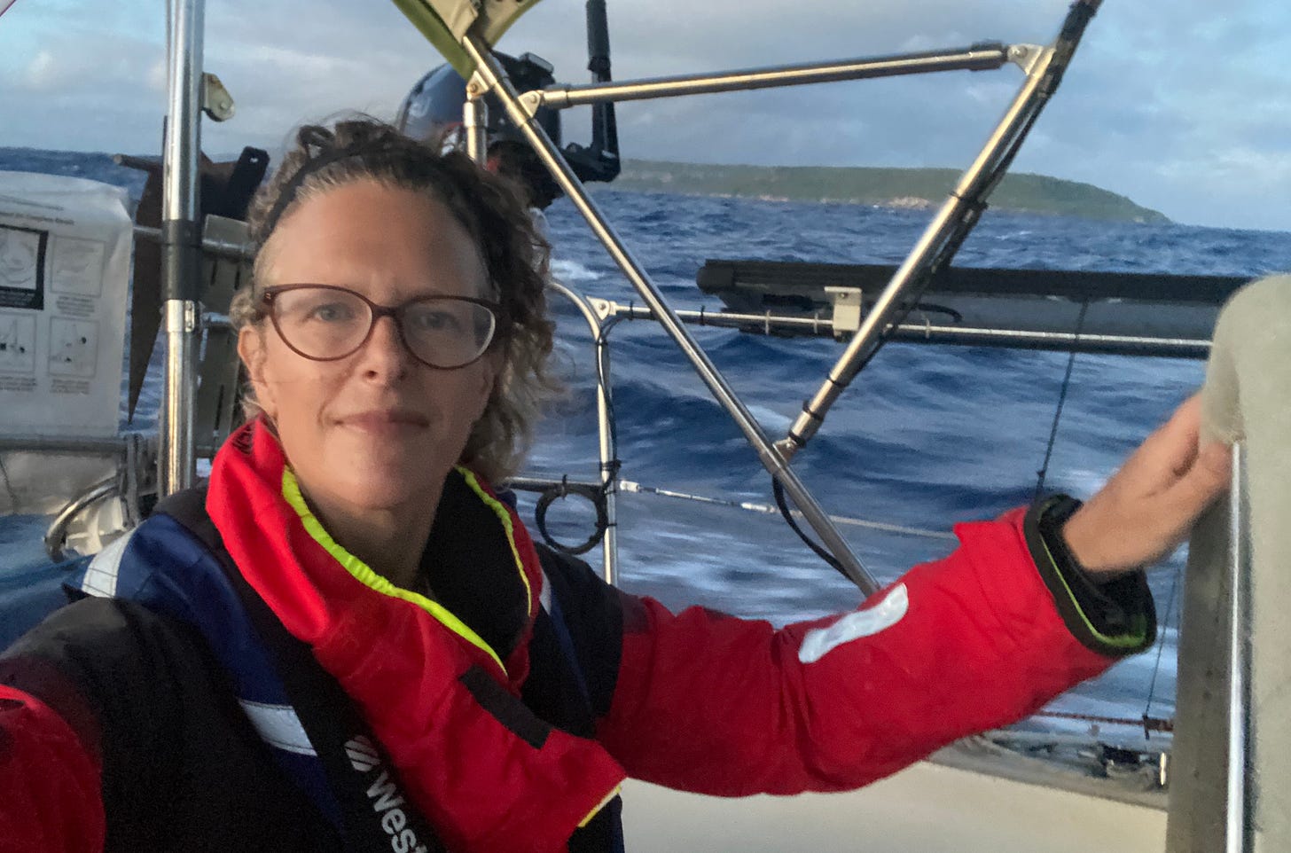 A woman takes a selfie from the cockpit of a sailboat in early morning light with land visible behind her