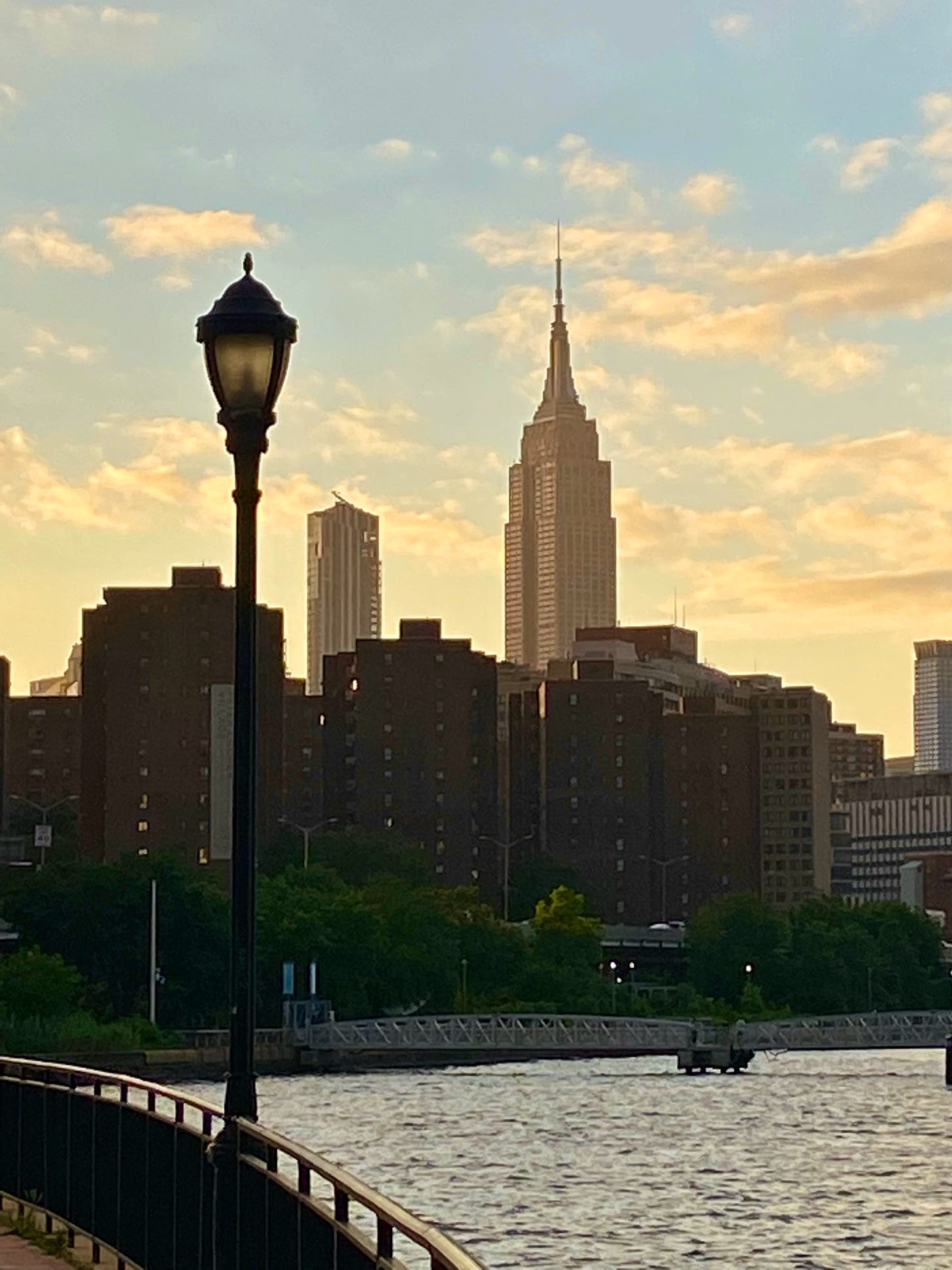 The Empire State Building seen from the East River. The sky is pale blue with glowing clouds. In the foreground is a black lamppost.