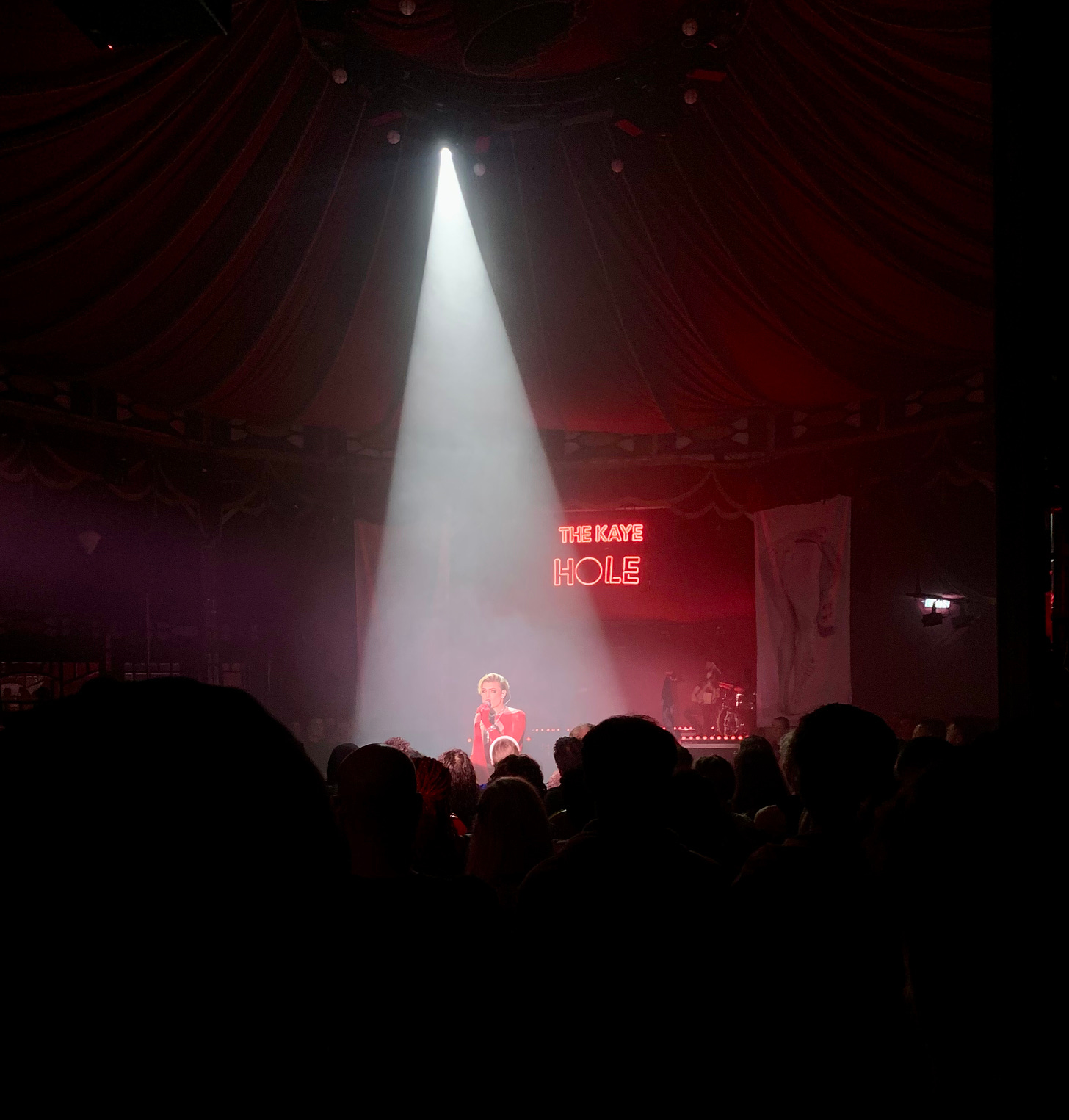 performer stands under a shaft of light on stage with a mic. There is a big audience watching and "The Kaye Hole" written in lights behind