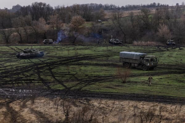 Tanks and other military vehicles conduct test-fire exercises in a valley marked by the many tank and tire tracks on the grass.