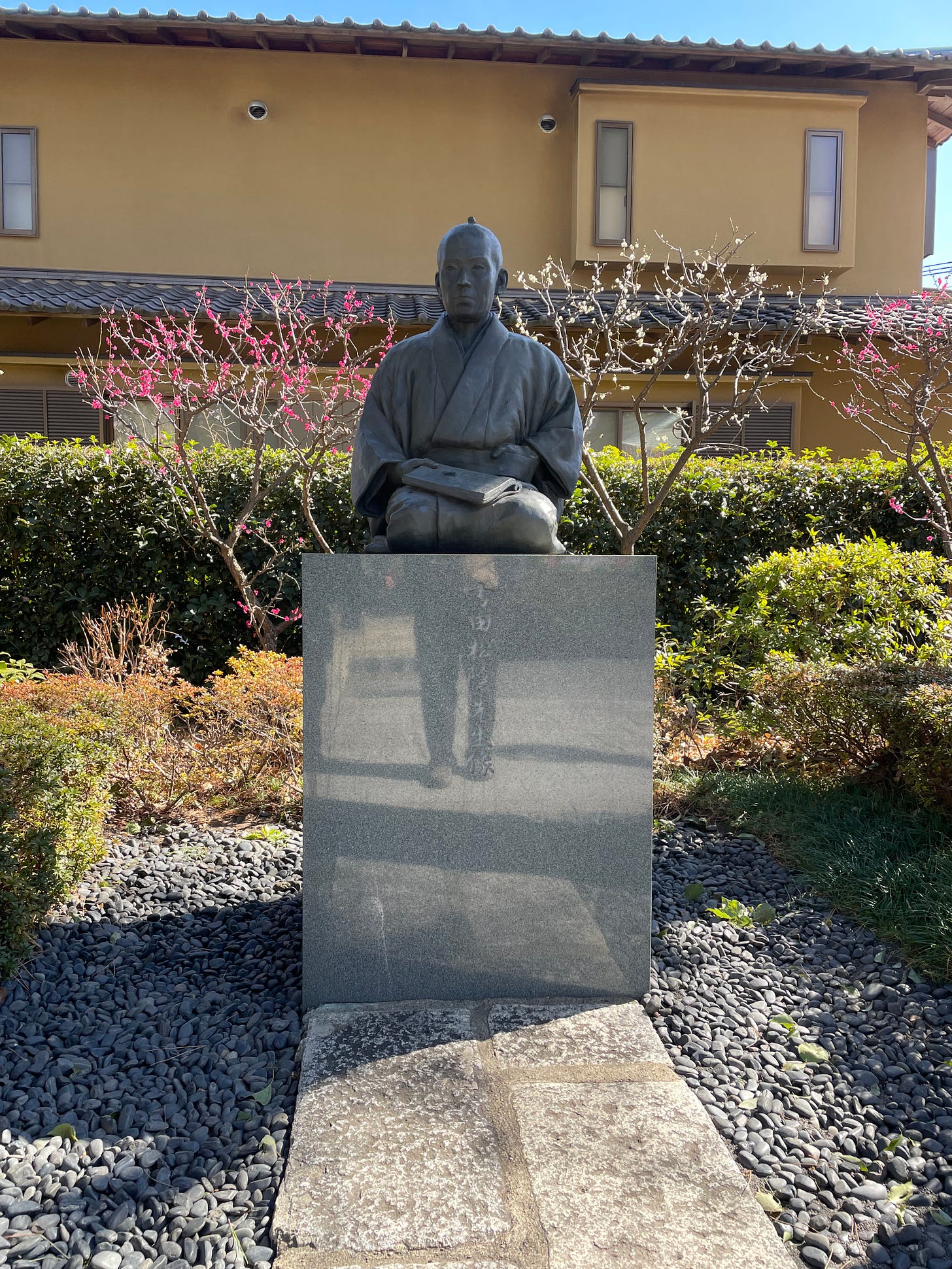 Statue of Shoin-san. In the background, plum trees are blooming in with pink and white buds.