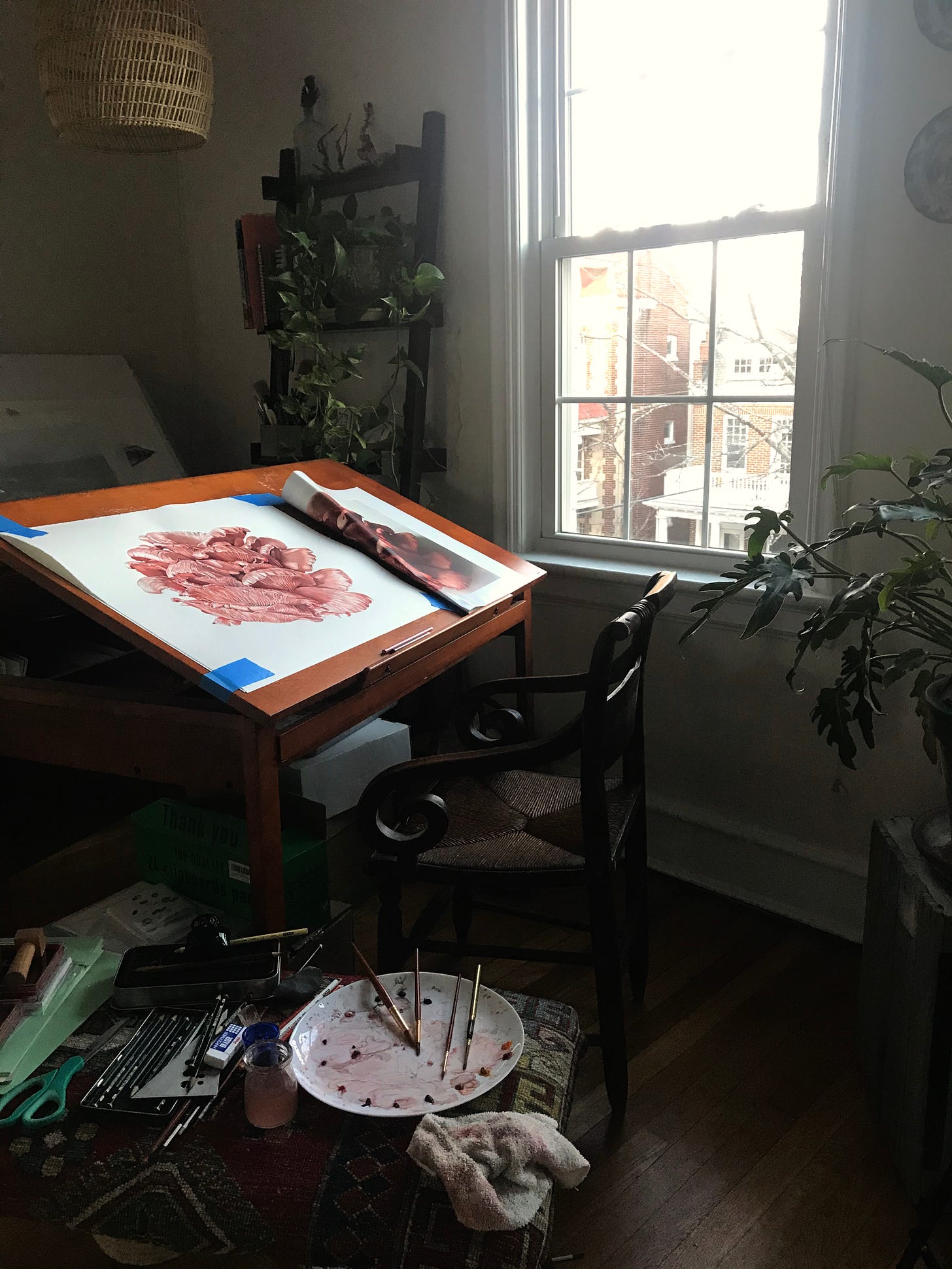 A photo of the studio space, the finished painting taped to the desk, with a window and plants close by