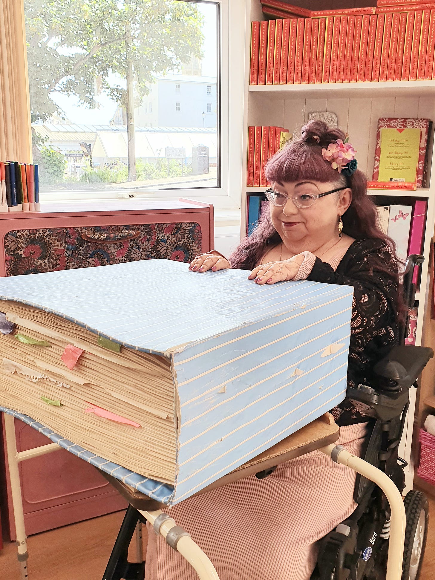 A woman with long red hair and glasses is sitting in front of a bookcase in a wheelchair. She is smiling and resting her hands on a very large oversized book