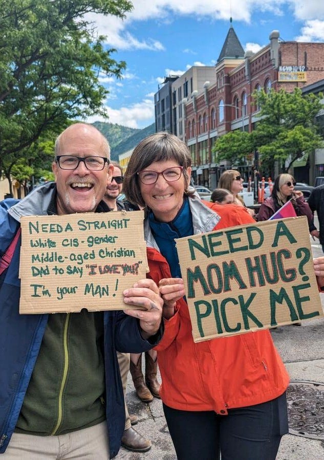 picture of two middle-age people in raincoats along the side of a street with other people standing around them. The man's cardboard sign says "Need a straight white cis-gendered middle-aged Christian dad to say I love you? I'm your man." The woman's sign says "Need a mom hug? Pick me." Both people are smiling broadly.