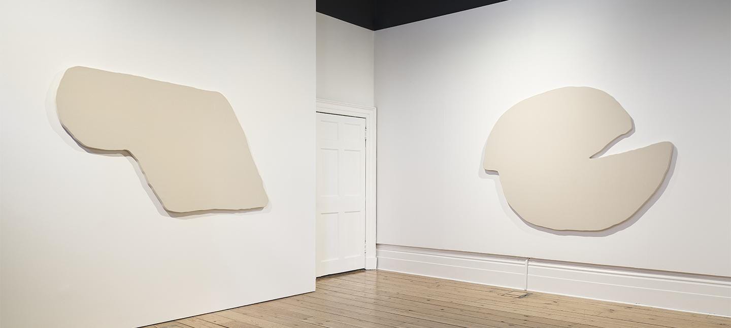An installation view of Christine Sun Kim's work in Gallery 31. Two large stretched canvases in abstract shapes hand either side of a door.