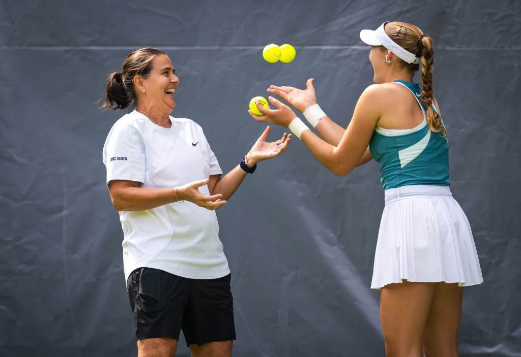 A young female tennis player wearing a teal tank top, white skirt, and white visor playfully juggles tennis balls with her coach, who is dressed in a white Nike shirt and black shorts. Both are smiling and laughing, sharing a lighthearted moment during a training session. The background features a simple grey tarp, likely at a practice court.