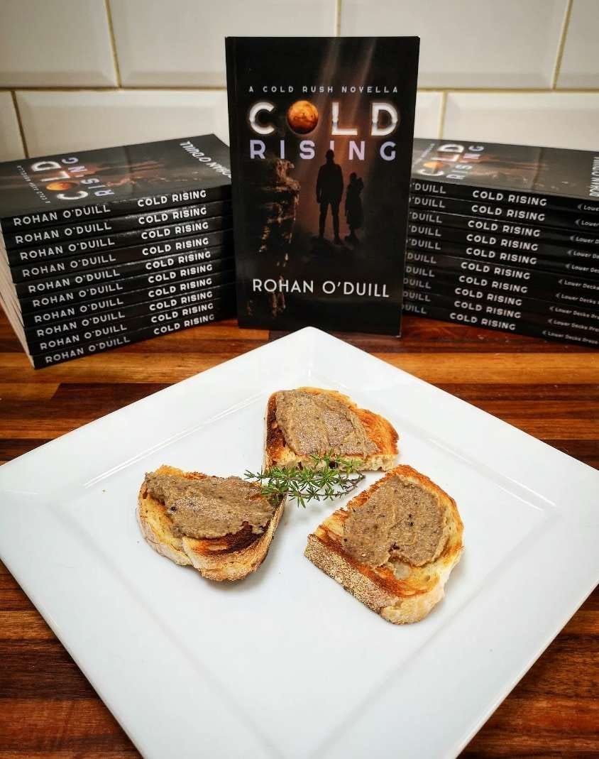 Cold Rising, a Cold Rush Novella by Rohan O'Duill behind a plate of bread with a mushroom spread on it. Yum!