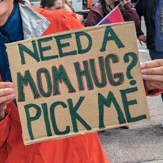 a close up of a woman in a red raincoat holding a cardboard sign that says, "Need a mom hug? Pick me."