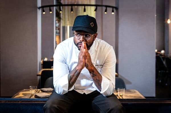 A chef in a white jacket and blue cap presses his hands together.