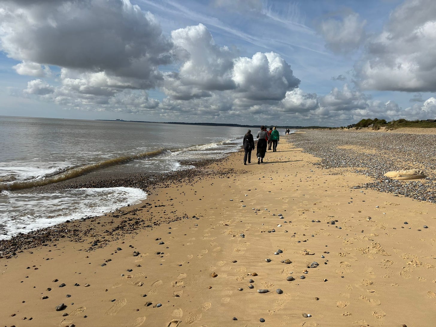 A photo of writers and researchers walking on a beach in Suffolk, talking about their research. The beach has pebbles on it, the waves lap gently at the shore, and the clouds look dramatic.