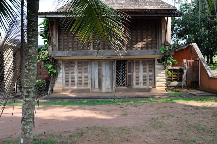 A two-storey house with large wooden doors and the entire front comprised of slatted wooden shutters in a yard with a palm tree.