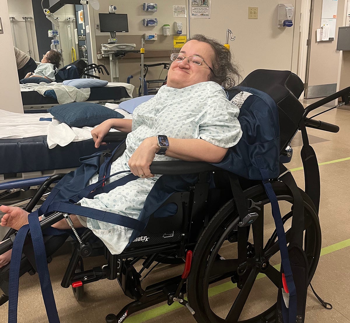 Athena in a hospital gown sitting in a manual wheelchair. She is straining to pull herself up with the armrests and smiles at the camera.