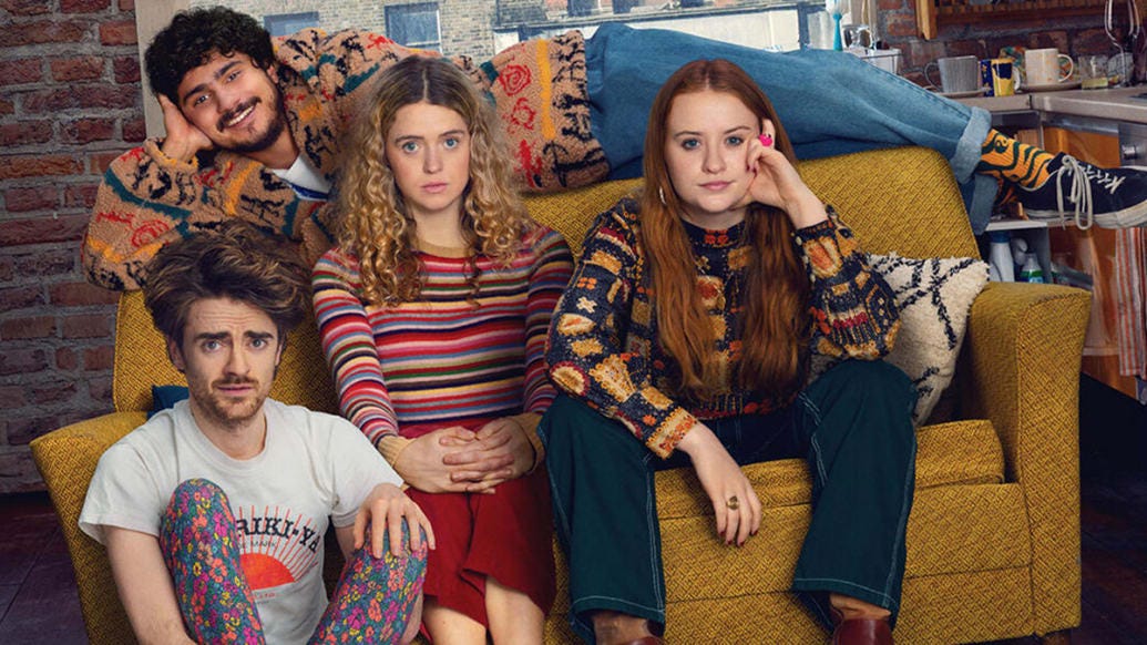Four actors (Luke Rollason, a white man with wavy brown hair and facial hair wearing a white t-shirt and bright floral print leggings; Bilal Hasna, a British-Palestinian man with curly black hair and facial hair wearing a patterned sweater and jeans; Sofia Oxenham, a white woman with long curly blond hair wearing a colorful striped shirt and a red skirt; and Máiréad Tyers, a white woman with long straight red hair wearing a patterned sweater and jeans) sit on a yellow couch in an apartment in a promo image for the Disney+ series Extraordinary. 