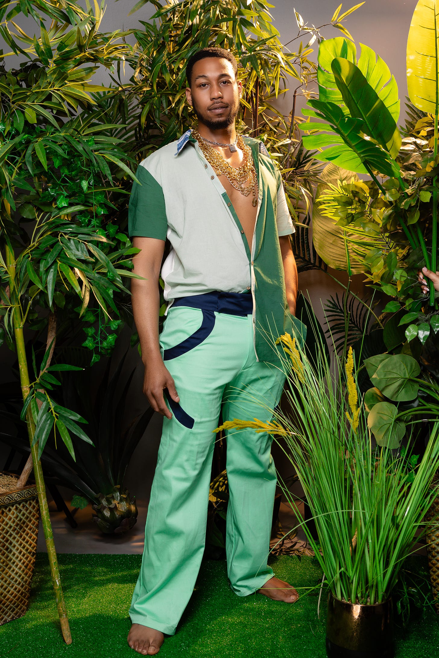 A man stands in a jungle-like setting wearing an outfit with a variation of greens and blues.