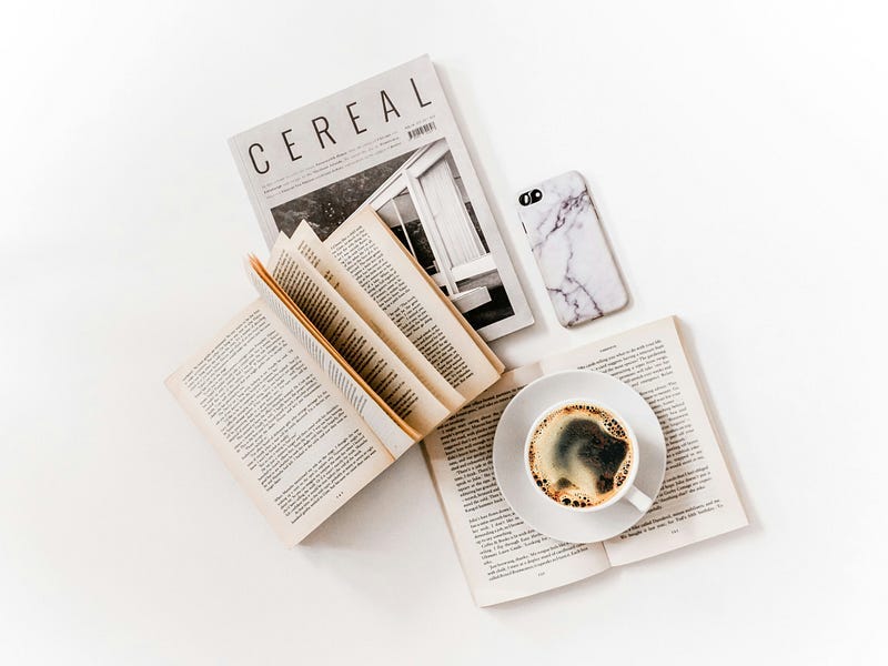 A cup of coffee on a book, on a table with other items like a book, magazine and a mobile. A cup of coffee on a book, on a table with other items like a book, magazine and a mobile.