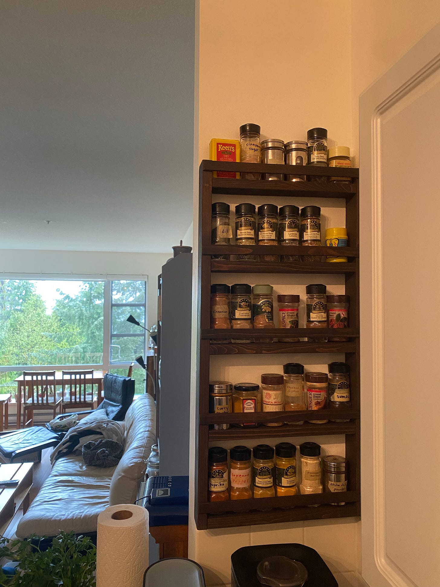 A section of kitchen wall with a wood spice rack mounted on it, with five shelves of various shapes and sizes of spice containers. In the background the couch and dining table are visible.